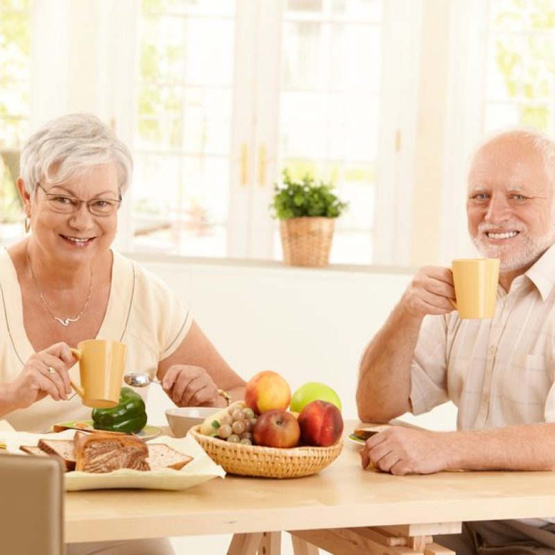Senior couple having tea at breakfast, sitting in sunny kitchen, smiling.